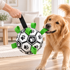 A golden retriever dog holds a black soccer ball toy with green loops in its mouth while a person's hand lifts the black handle. The toy features black paw prints on a white background, set in a bright, modern living room.