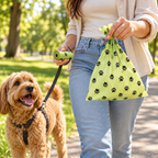 A woman in a park holds a green plastic bag with black paw print patterns and a matching green leash holder, with a small brown dog on a black leash beside her.